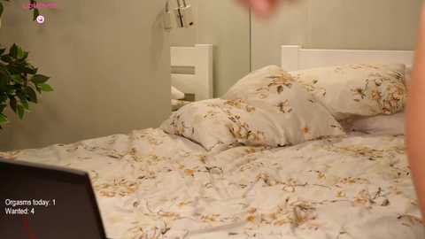 Media: Video of a bedroom with a white bed adorned with a floral-patterned duvet and pillows, beige walls, and a potted plant in the corner.