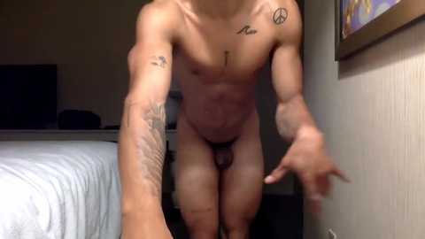 Media: Video of a fit, nude man with a small penis and tattoos, standing in a dimly lit room with a bed and framed artwork.