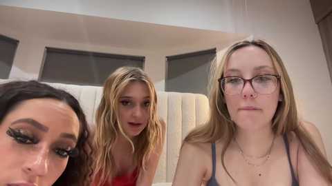 Media: Video of three young women, two with blonde hair, one with dark hair, seated in a modern, light-colored room with white cushions and windows.