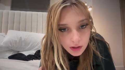 Media: A video of a blonde, fair-skinned woman with long hair, wearing black clothing, leaning over a bed with white linens. The background features a modern, minimalist bedroom with a white headboard and a warm, soft light source.