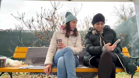 Media: Video of two women, one in a green beanie and puffy coat, the other in a black beanie and puffer jacket, sitting on a wooden bench in a cold, barren garden.