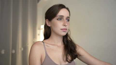 Media: Video of a young Caucasian woman with fair skin and long, dark brown hair, wearing a beige tank top, standing in a modern, neutral-toned room with minimalistic decor.