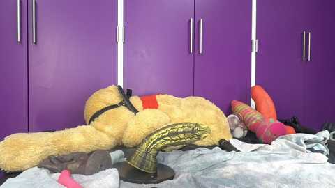 Media: Video of a plush astronaut teddy bear suspended by white cords in a purple wardrobe, surrounded by colorful, patterned toys on a messy bed.