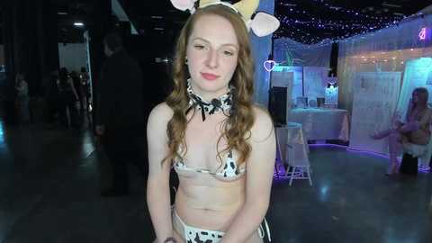 Media: Video of a fair-skinned, red-haired woman wearing a cow-themed lingerie set, posing indoors at a dimly lit, festive event with string lights and a DJ booth in the background.