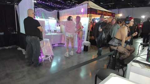 Media: Video of a crowded, dimly lit indoor event space with various vendors and attendees. Booths display colorful merchandise, including lingerie and costumes. People, mostly women, in revealing outfits and lingerie are seated or standing, some wearing headphones.