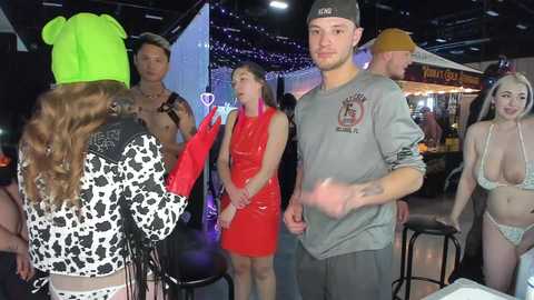 Media: Video of a lively indoor bar scene with a tattooed man in a cap, a woman in a leopard print top, and others in bikinis and casual attire, all smiling and interacting.