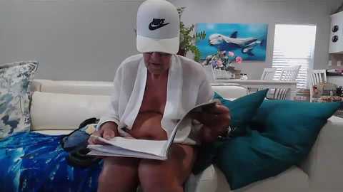 Media: Video of a shirtless, middle-aged man with a white cap, reading a book on a white sofa in a modern living room with blue cushions, white walls, and a dolphin painting.