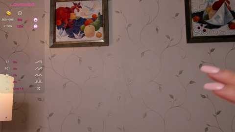 Media: Video of a beige wallpapered room with abstract fruit paintings, a small white lamp, and a hand with manicured nails holding a controller.