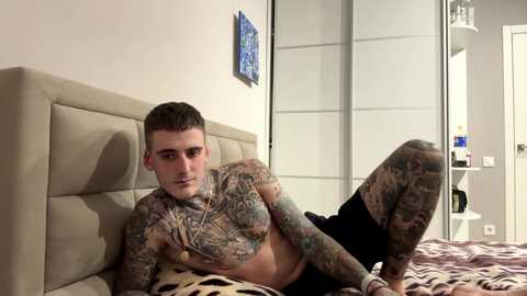 Media: Video of a tattooed, shirtless, fair-skinned man with short dark hair lying on a bed with zebra-patterned sheets, in a modern bedroom with white walls and a sliding wardrobe.