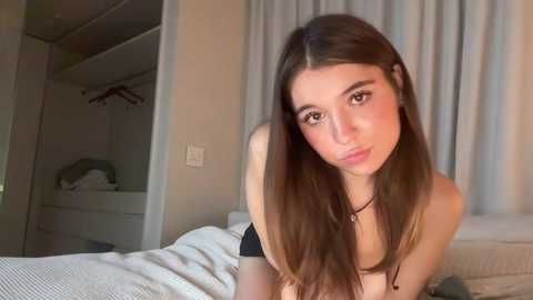 Media: A video of a young woman with long, straight brown hair, light skin, and brown eyes, wearing a black top, leaning forward on a bed with white sheets and a gray cushion. Background features a closet and beige curtains.