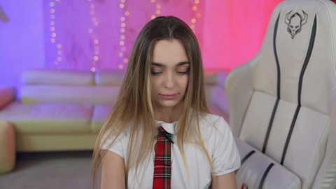 Media: Video of a young woman with long brown hair, wearing a white blouse and a red plaid tie, sitting in a white gaming chair with a skull logo. Background features a pink and purple string light ambiance and a beige couch.