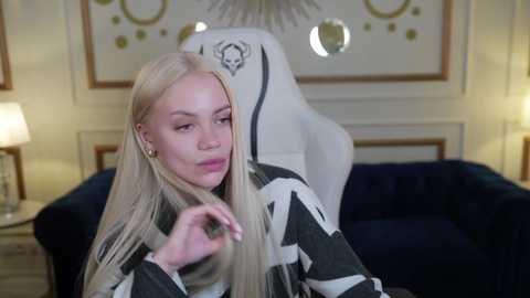 Media: Video of a blonde woman with long hair, wearing a black and white striped sweater, adjusting her hair, sitting in a white gaming chair in a modern, dimly lit room with a blue sofa.