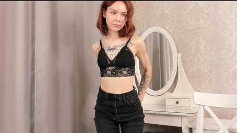 Media: Video of a slender, fair-skinned woman with red hair, wearing a black lace bra and high-waisted jeans, standing in a room with a white vanity and floral wallpaper.