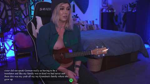 Media: Video of a blonde woman with medium-sized breasts, wearing a green shirt, playing a ukelele in a cozy, dimly-lit room with a bed, plants, and a gaming chair.