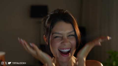 Media: Video of a joyful woman with dark hair, open mouth, and raised hands, in a dimly lit room, with a watermark in the bottom left corner.