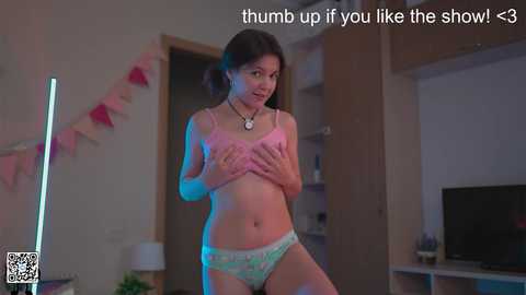 Media: A video of a young Asian woman with fair skin, dark hair, and small breasts, wearing a pink bra and light blue panties, covering her chest with her hands in a cozy, dimly lit room.