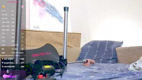 Media: Video of a modern bedroom with a gray bedspread, beige headboard, and a black exercise bike with a digital display. A person is lying on the bed with a hand holding a small device.