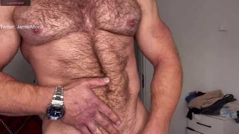 Media: Video of a muscular, hairy-chested man, wearing a silver wristwatch, standing in a bedroom with a bed, clothes on a dresser, and a white wall.