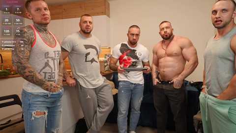 Media: Video of four shirtless, muscular men in casual clothes, one with tattoos, standing in a dimly lit room with a wooden ceiling and shelves in the background.