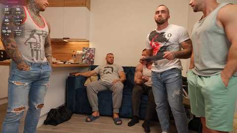 Media: Video of four muscular men in casual attire; one man in a tank top with tattoos, another in a graphic T-shirt, and two in light green shorts, interacting in a modern, minimalist living room with wooden accents.