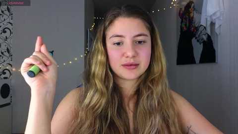 Media: Video of a young woman with long, wavy, light brown hair, fair skin, and a slim build, holding a green nail polish bottle, standing in a cozy room with string lights, black and white art on the wall, and a white garment hanging.