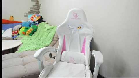 Media: Video of a white gaming chair with pink accents, next to a green crocheted blanket on a beige sofa, with colorful stuffed toys and a bookshelf in the background.