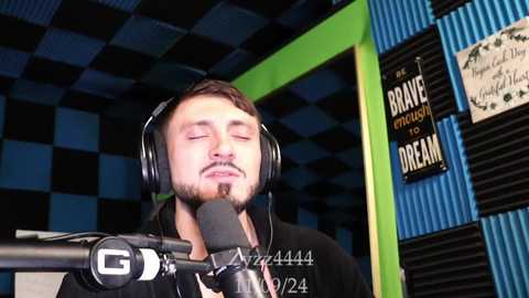 Media: Video of a bearded man in a black shirt singing into a microphone against a blue-and-green checkered wall.