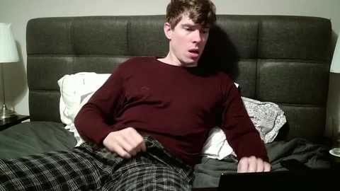 Media: Video of a young man with short brown hair, wearing a burgundy shirt and plaid pajama pants, sitting on a bed with a gray headboard. He looks surprised, holding a laptop.