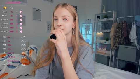 Media: A video of a young woman with long blonde hair, wearing a gray shirt, holding a phone, in a cluttered bedroom with a white bed, shelves, and clothes.