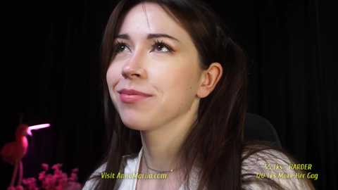 Media: Video of a fair-skinned woman with long brown hair, wearing a white shirt, smiling slightly. Background is dark, with a pink light and text overlaid.