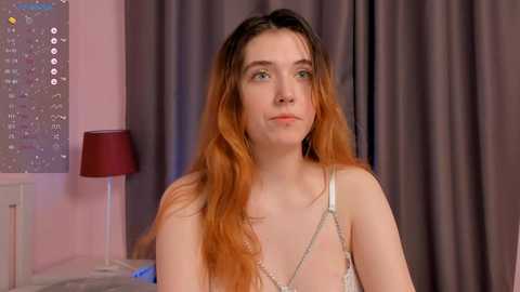 Media: Video of a fair-skinned young woman with long, wavy, auburn hair, wearing a sheer white bralette. She has a slender build and is seated in a cozy room with pink walls, a red lamp, and gray curtains.