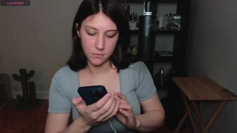 Media: Video of a young woman with shoulder-length dark hair, wearing a light blue t-shirt, intently looking at her smartphone in a dimly lit room with a wooden table and a shelf with various items in the background.