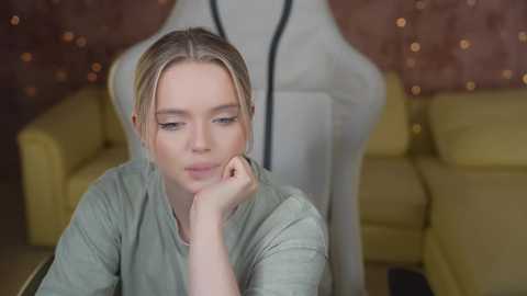 Media: Video of a young, fair-skinned woman with blonde hair, wearing a light green shirt, resting her chin on her hand, seated in a cozy room with yellow couches and a mannequin in the background.