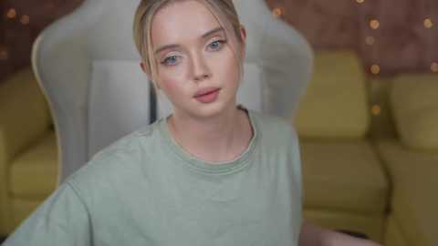 Media: A video of a young woman with fair skin, light blonde hair in a bun, and light green eyes, wearing a loose, mint-green top. She sits in a white chair with a yellow cushion, against a backdrop of a cozy living room with warm, bohemian-style decor.