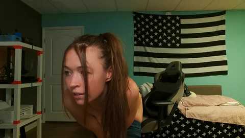 Media: A video of a young woman with long brown hair in pigtails, leaning forward in a cluttered bedroom with a gaming chair and an American flag wall hanging.