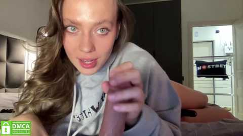 Media: Video of a young, fair-skinned woman with long, wavy brown hair, wearing a gray hoodie, lying on a bed, looking shocked with her hands clenched, in a modern bedroom with a dark headboard and white furniture.