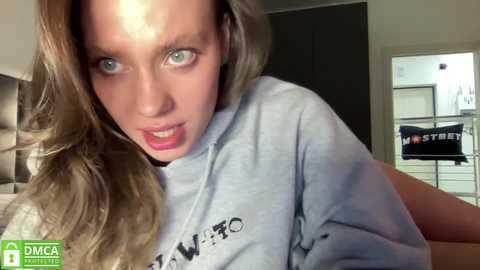 Media: A video of a young woman with long blonde hair and striking blue eyes, wearing a gray sweatshirt, looking directly at the camera with a slightly intense expression. Background features a bedroom with a bed and white wardrobe.