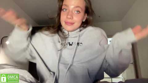 Media: A video of a young woman with light skin and brown hair, wearing a grey hoodie, smiling and waving. Background shows a bedroom with white walls and a bed.