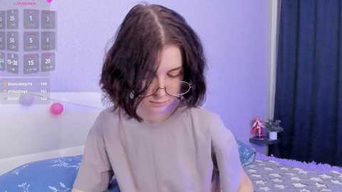 Media: Video of a young woman with shoulder-length dark hair, wearing glasses and a gray shirt, sitting on a bed with a blue and white patterned blanket. Background includes a white wall with a periodic table poster and a pink ball.