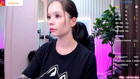 Media: Video of a young Asian woman with fair skin and dark hair, wearing a black t-shirt, seated in a dimly-lit room with green plants, a chair, and streaming setup visible.