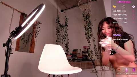 Media: Video of a young Asian woman with long black hair, topless, sitting in a white chair in a dimly lit room, holding a smartphone. Background shows a studio with a ring light, a desk, and hanging plants.