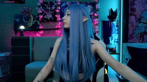 Media: A video of a young woman with long, straight, blue hair, dressed in a black leather outfit, sitting in a dimly lit room with neon lights and a floral wall mural.