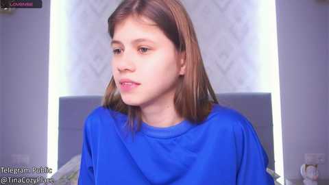 Media: Video of a young Caucasian woman with straight, shoulder-length brown hair, wearing a blue shirt, sitting in a modern bedroom with a grey headboard and patterned wallpaper.