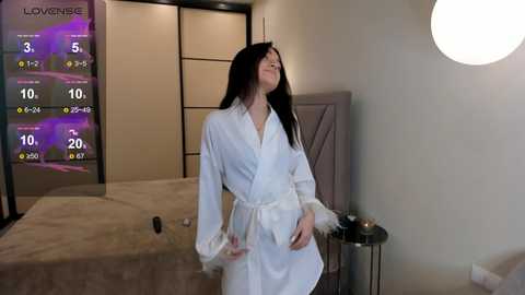 Media: Video of a woman in a white robe with fur trim, standing in a modern bedroom with a bed, a TV displaying weather, and a round light fixture.