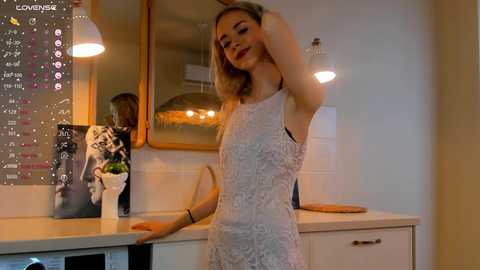 Media: Video of a blonde woman in a white lace dress, standing in a modern kitchen with a calendar, plants, and warm lighting.