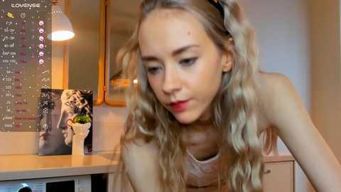 Media: Video of a young Caucasian woman with long, wavy blonde hair, wearing a sleeveless top, leaning over a desk. Background includes a calendar, a potted plant, and a mirror.