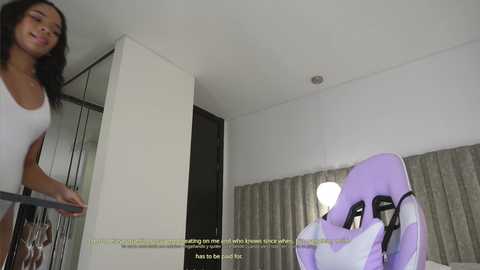 Media: Video of a woman with long dark hair in a white tank top, standing in a modern, minimalist bedroom with white walls, a purple gaming chair, and beige curtains.