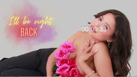 Media: Video of an Asian woman with long black hair, wearing a strapless black dress adorned with pink roses, lying on a bed. Background features a pastel gradient and text \"I'll be right BACK\" in yellow and red.