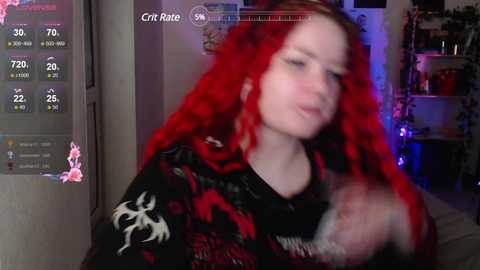 Media: Video of a young woman with long, vibrant red hair, wearing a black shirt with red and white patterns, playing a video game. Background shows a dimly lit room with a TV displaying game stats.