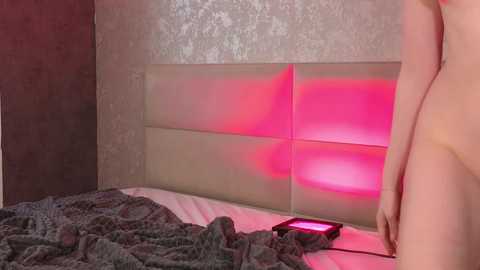 Media: Video of a nude, light-skinned person standing beside a bed with a gray, textured blanket and a pink light glowing on a headboard.